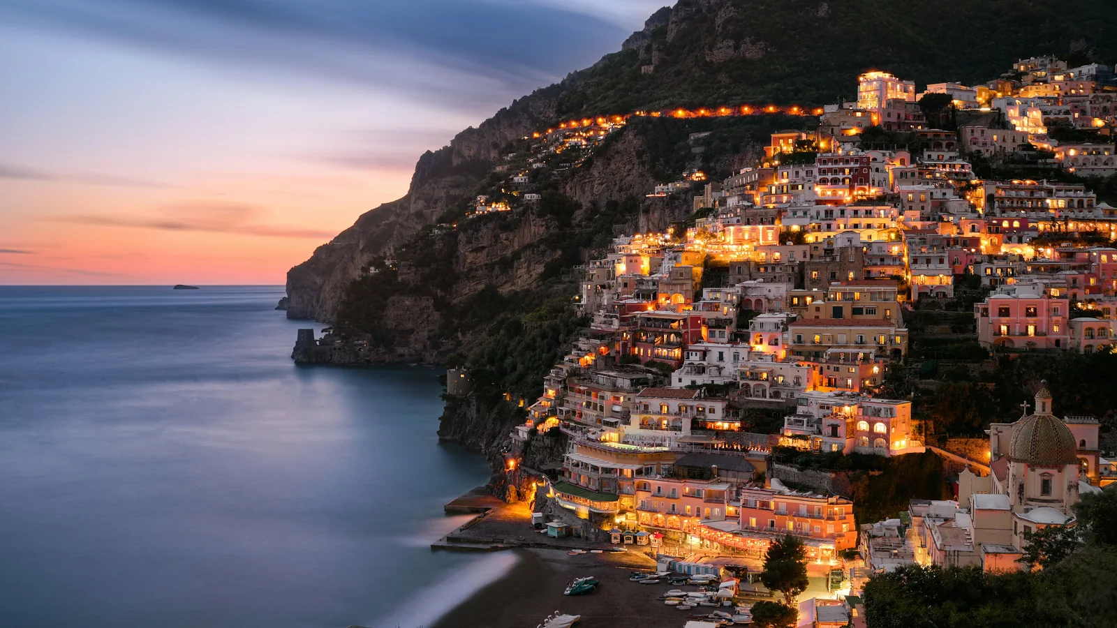 Positano's houses stacked in tiers up the cliff, lit at dusk above the beach
