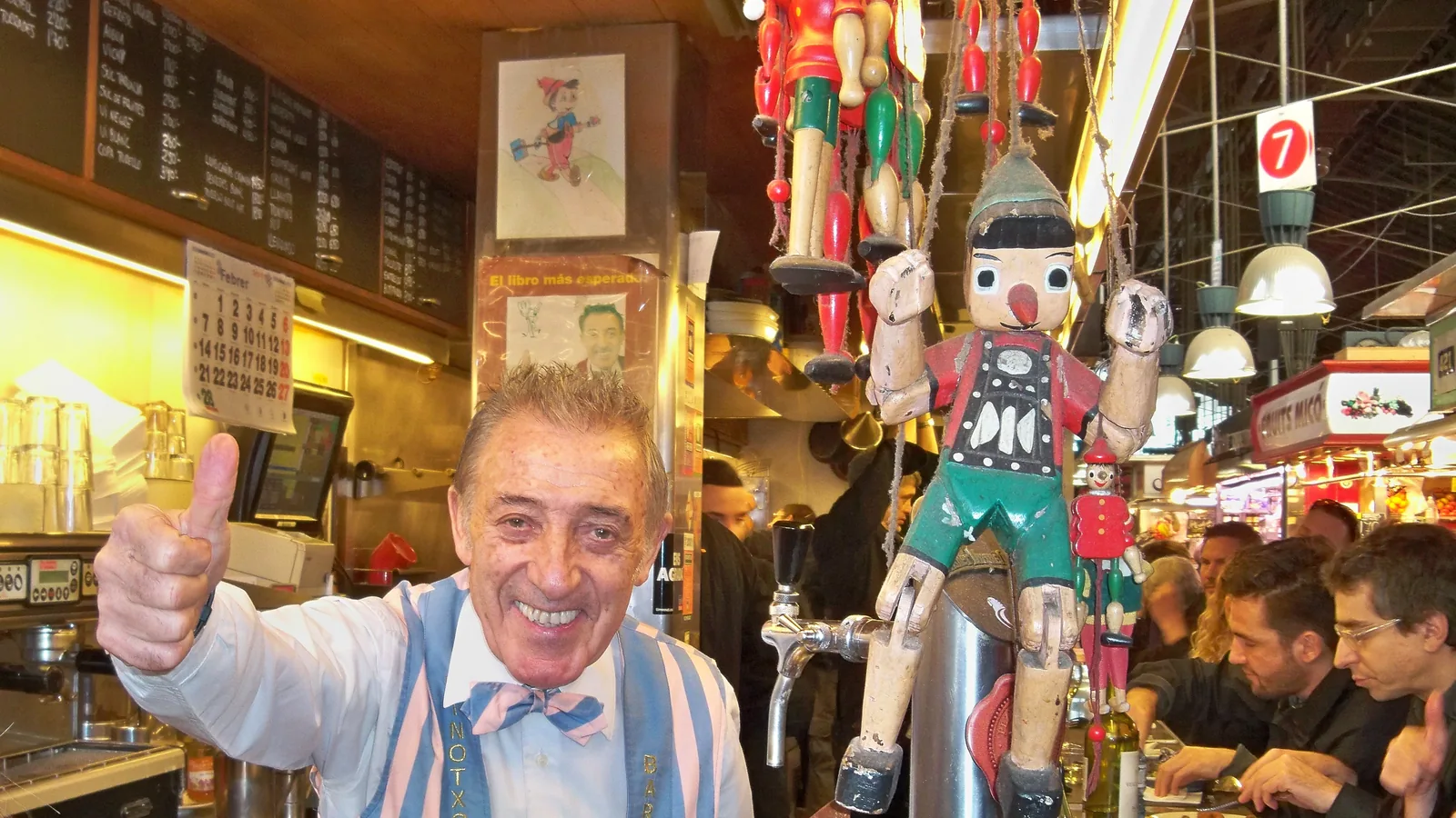 The counter of Bar Pinotxo at its original stall inside Mercat de la Boqueria, with the barman in the bar's embroidered striped waistcoat, chalked menu boards and customers eating at the bar