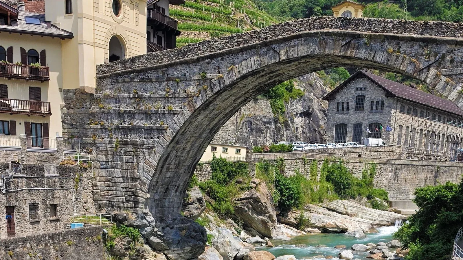 An old stone arch bridge spanning a rocky river below a hillside village.