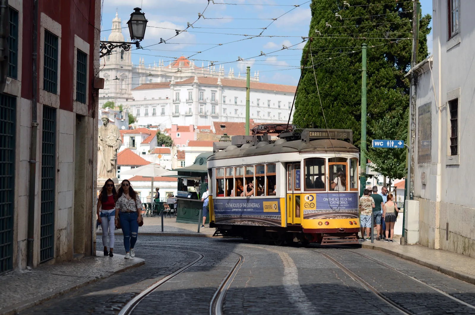 A yellow Lisbon tram on route 28E on the cobbled street at Largo de Santa Luzia in Alfama, with the Monastery of São Vicente de Fora on the skyline behind