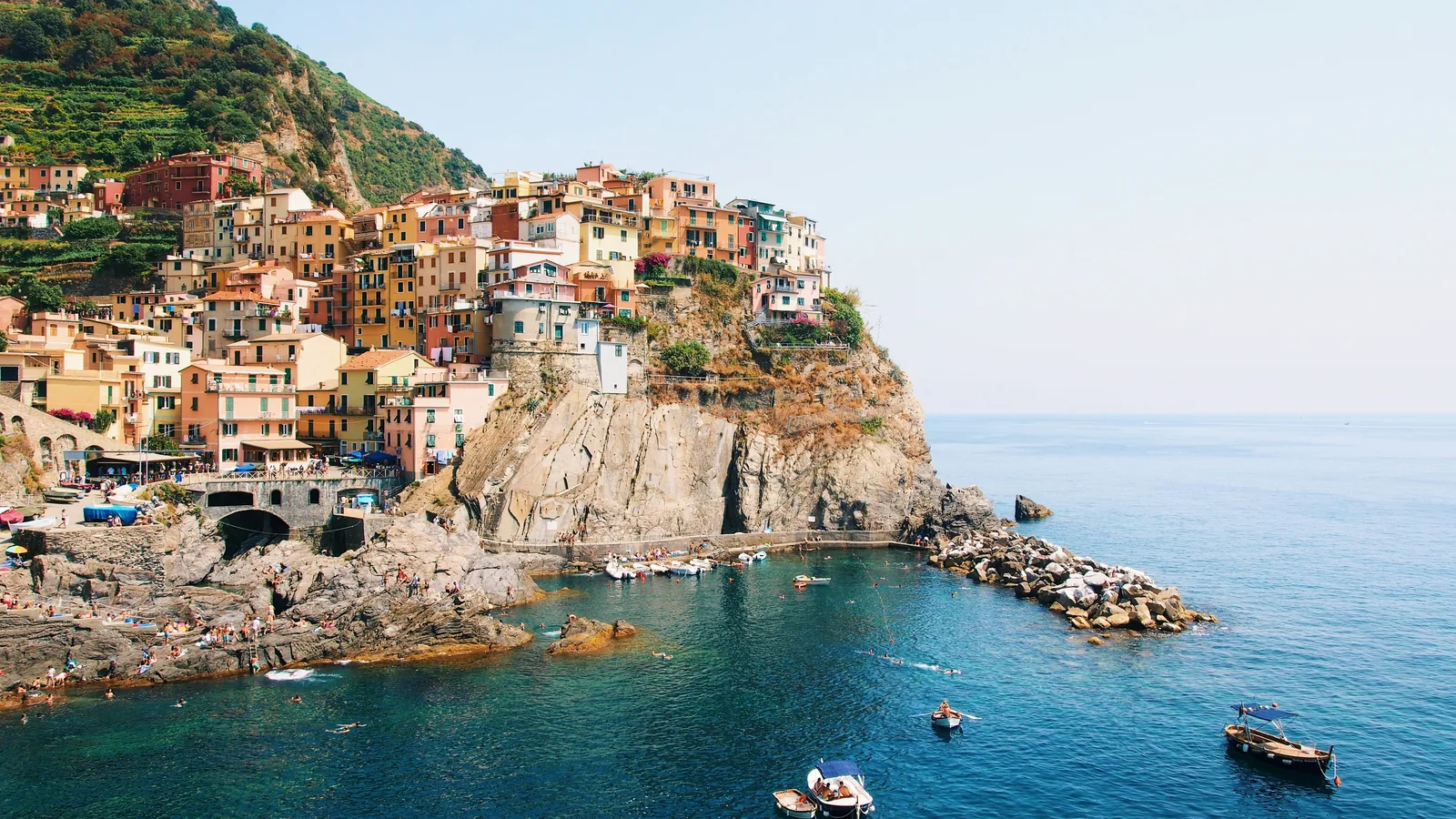 Manarola's pastel houses stacked on a rocky headland above a small harbour with swimmers and boats, Cinque Terre