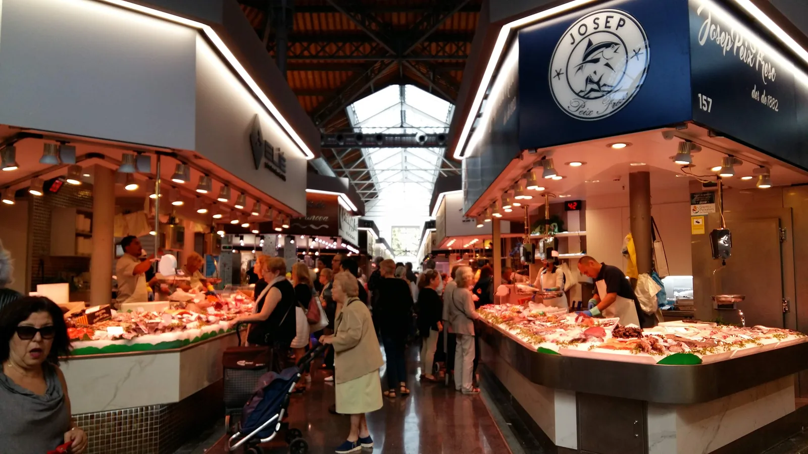 The central aisle of the renovated Mercat de Sant Antoni in Barcelona, between two fresh fish counters, with shoppers queueing under the market's iron roof trusses and skylight
