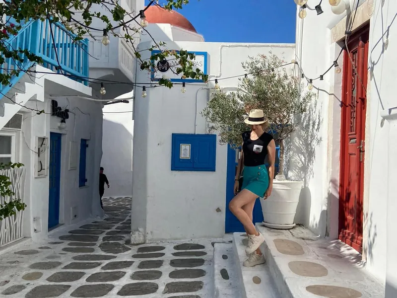 The writer standing on the steps of a whitewashed Cycladic lane in a straw hat, with festoon lights overhead, a red door beside her and a red-domed church behind